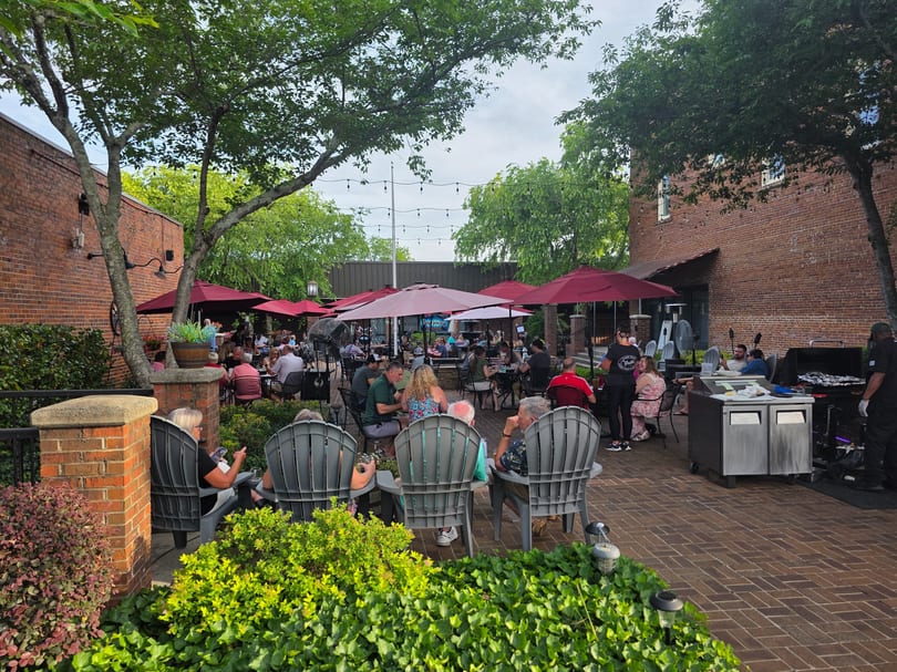 Patrons dine outdoors on a brick patio under red umbrellas and string lights, surrounded by lush greenery.