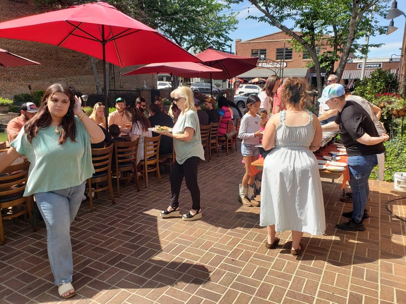People gather on a sunny outdoor patio with red umbrellas, enjoying food and drinks.