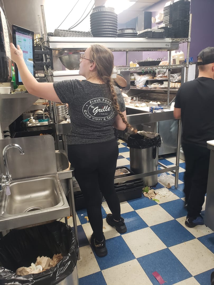 A woman in a "The Grille" shirt uses a touchscreen in a busy commercial kitchen.