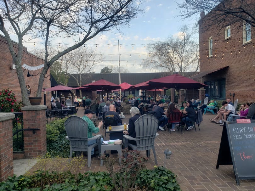 Patrons enjoying an outdoor patio with red umbrellas, string lights, and brick buildings.