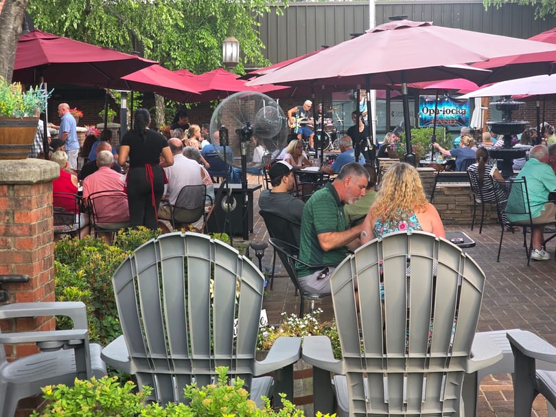 Patrons enjoying live music and dining under red umbrellas on a restaurant's outdoor patio.
