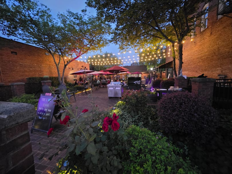 An outdoor restaurant patio at dusk, with string lights, red umbrellas, and lush greenery.