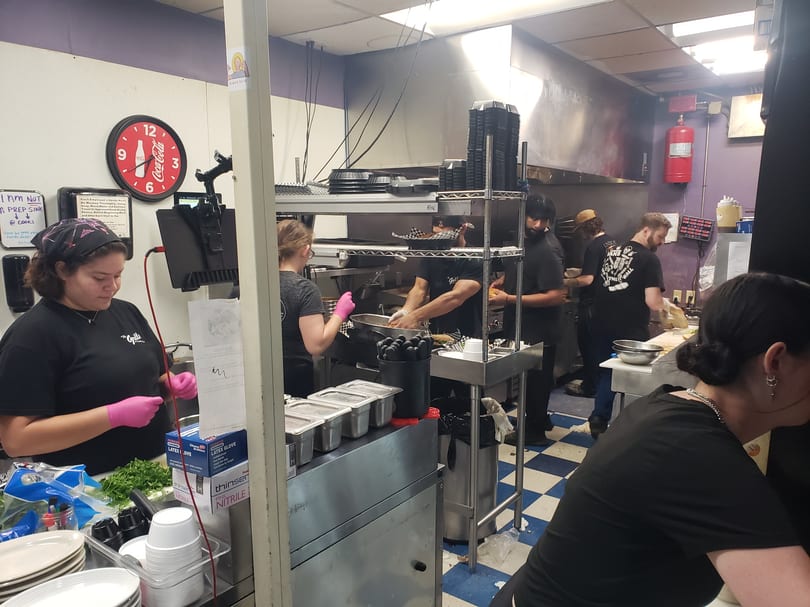 Busy restaurant kitchen with multiple cooks preparing food, stainless steel counters, and a checkered floor.
